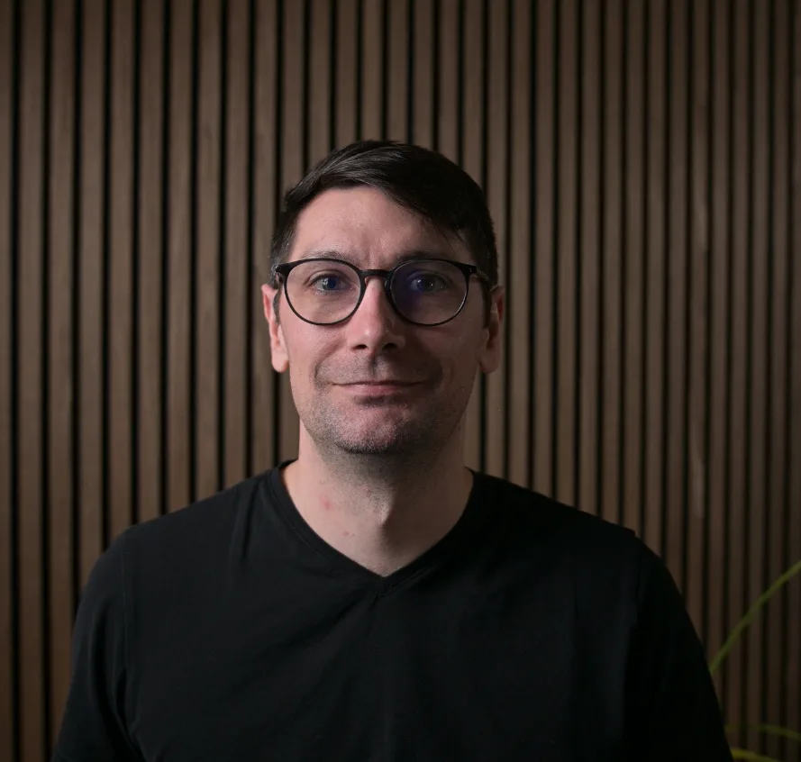 Portrait photo of an adult man wearing round black eyeglasses and a black V-neck shirt, facing the camera against a vertical wooden slat wall background. No readable text visible.